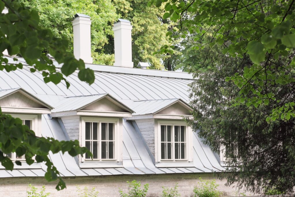 roof of an old house with chimney and attic roof windows surrounded by trees in summer.